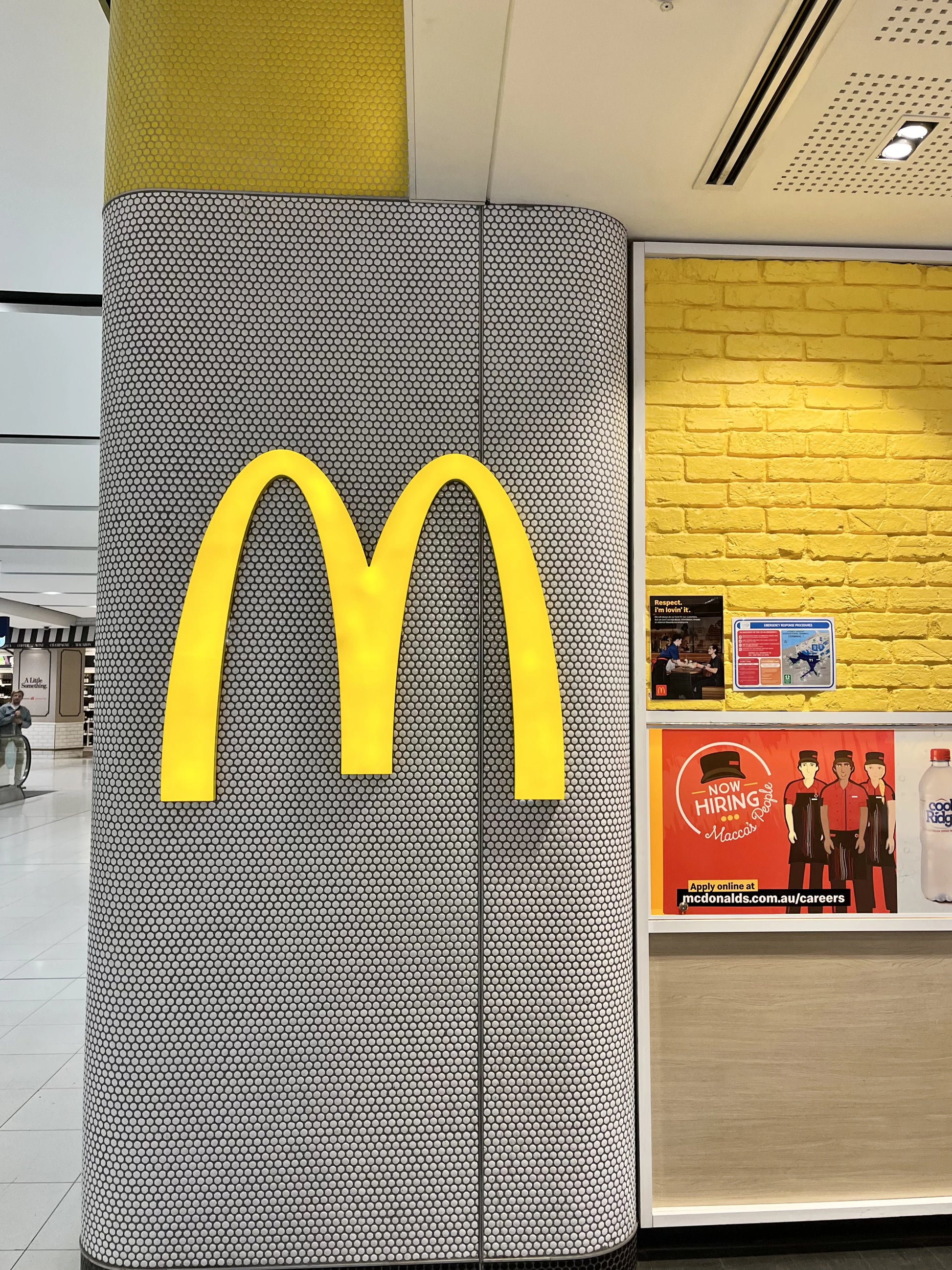 White Gloss Diameter Glazed Porcelain Penny Round 19mm tiles covering a large cylindrical column in a bright shopping centre interior with yellow accents.