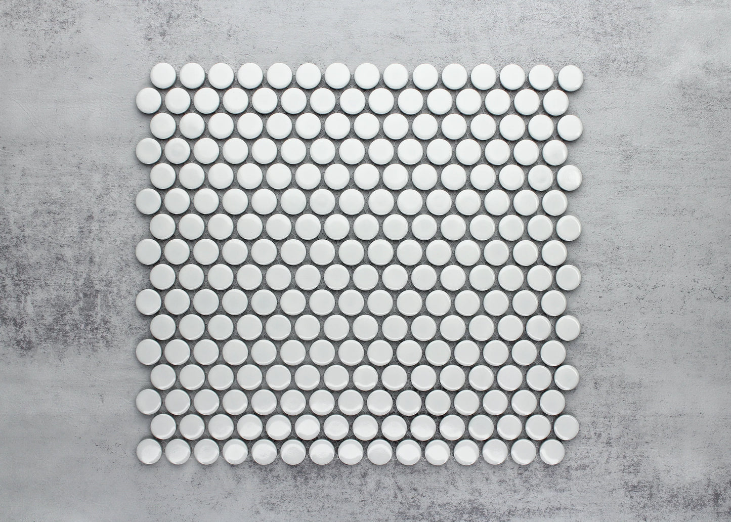 Flat lay pack shot of White Gloss Diameter Glazed Porcelain Penny Round 19mm tiles arranged in a uniform grid pattern on a grey concrete surface.