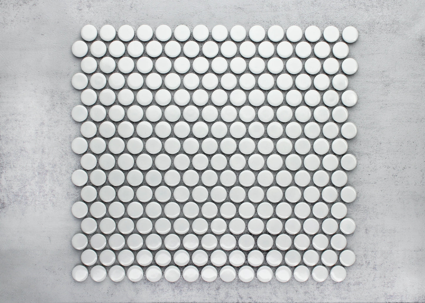Flat lay pack shot of White Gloss Diameter Glazed Porcelain Penny Round 19mm tiles arranged in a grid pattern on a grey concrete surface, showing their circular shape and glossy white finish.