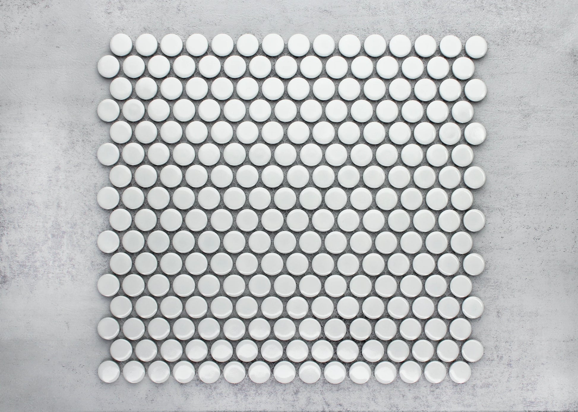 Flat lay pack shot of White Gloss Diameter Glazed Porcelain Penny Round 19mm tiles arranged in a grid pattern on a grey concrete surface, showing their circular shape and glossy white finish.