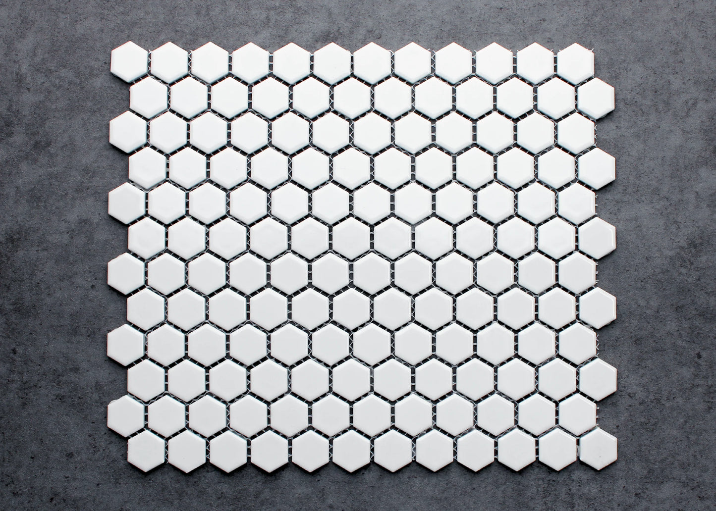 A flat-lay pack shot of White Gloss Porcelain Glazed Hexagon 23mm mosaic tiles arranged in a full sheet, showing glossy white hexagons with dark grout joints on a grey concrete background.