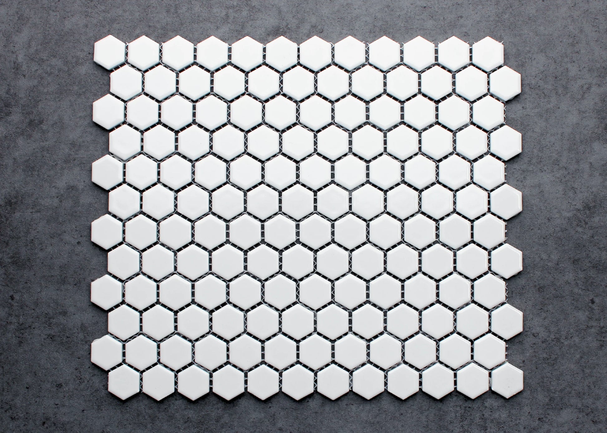 A flat-lay pack shot of White Gloss Porcelain Glazed Hexagon 23mm mosaic tiles arranged in a full sheet, showing glossy white hexagons with dark grout joints on a grey concrete background.