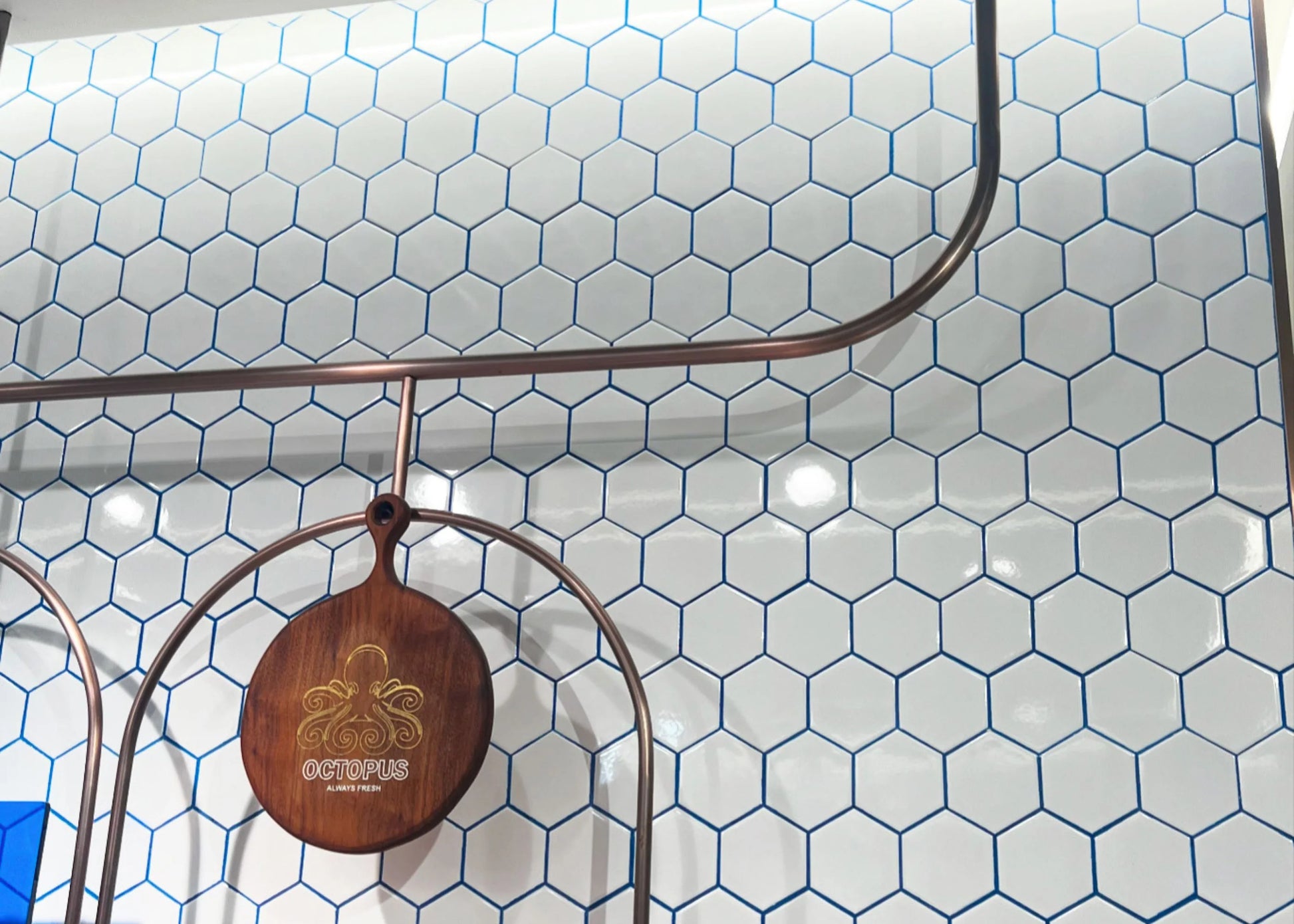 White Gloss Porcelain Glazed Hexagon 95x110mm tiles installed as a restaurant feature wall with blue grout lines, copper pipe railing, and a branded wooden Octopus sign.