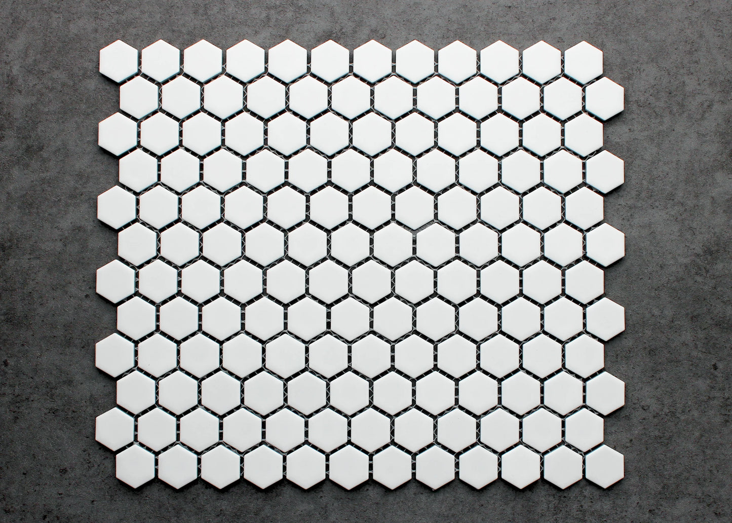 A flat-lay pack shot of White Matte Porcelain Glazed Hexagon 23mm tiles arranged in a honeycomb mosaic sheet pattern against a grey concrete surface.