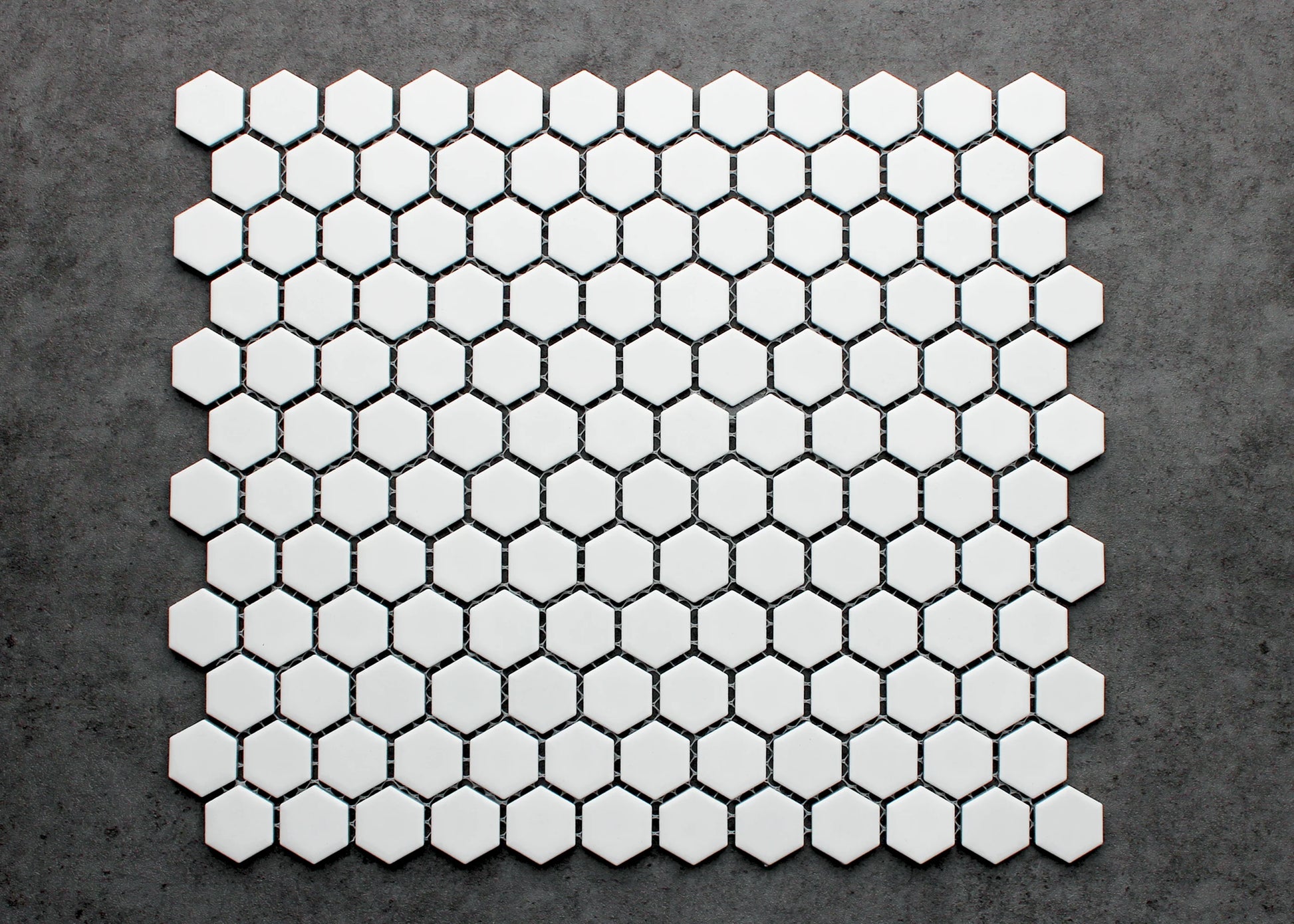 A flat-lay pack shot of White Matte Porcelain Glazed Hexagon 23mm tiles arranged in a honeycomb mosaic sheet pattern against a grey concrete surface.