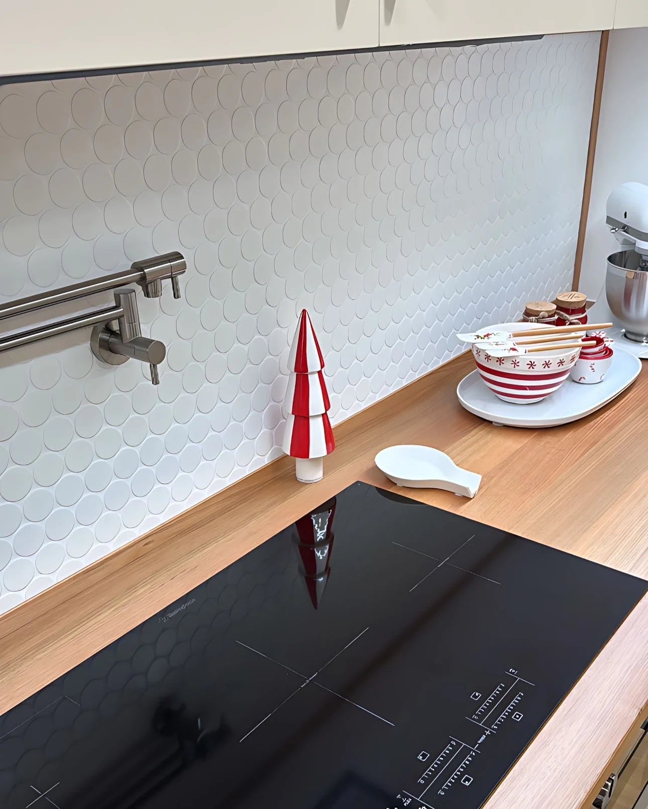 White Matte Porcelain Glazed Penny Round 48mm tiles installed as a kitchen splashback behind a black induction cooktop, pot filler faucet, and wooden benchtop with festive red and white accessories.