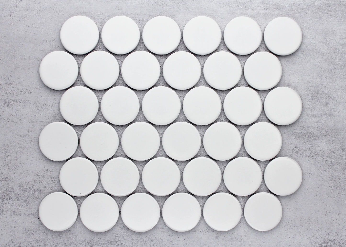 Overhead pack shot of White Matte Porcelain Glazed Penny Round 48mm tiles arranged in an offset grid pattern on a grey concrete surface.