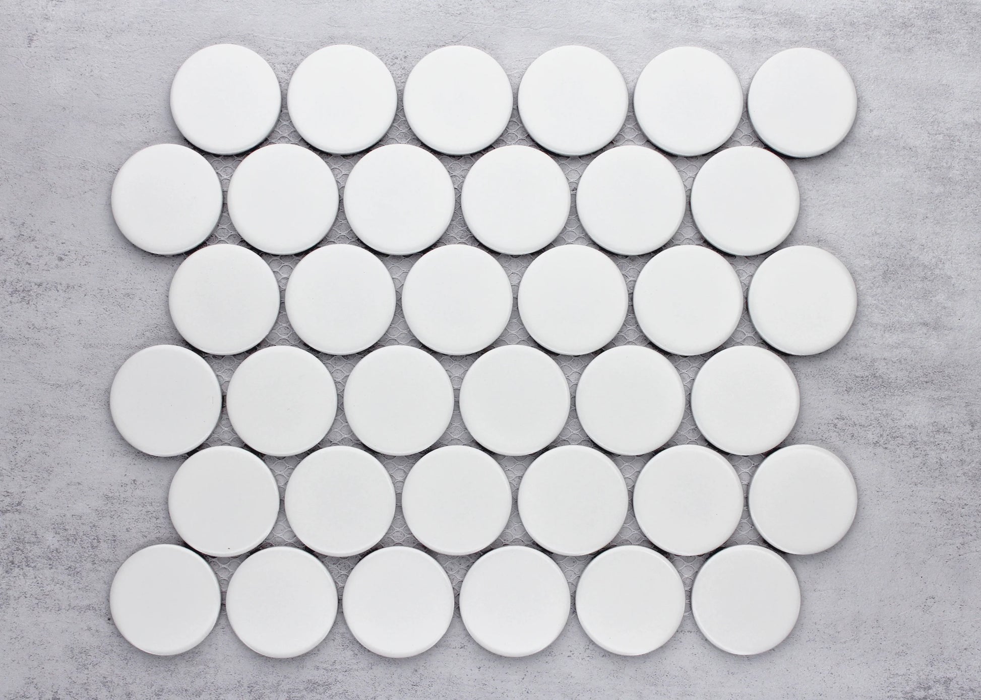 Overhead pack shot of White Matte Porcelain Glazed Penny Round 48mm tiles arranged in an offset grid pattern on a grey concrete surface.