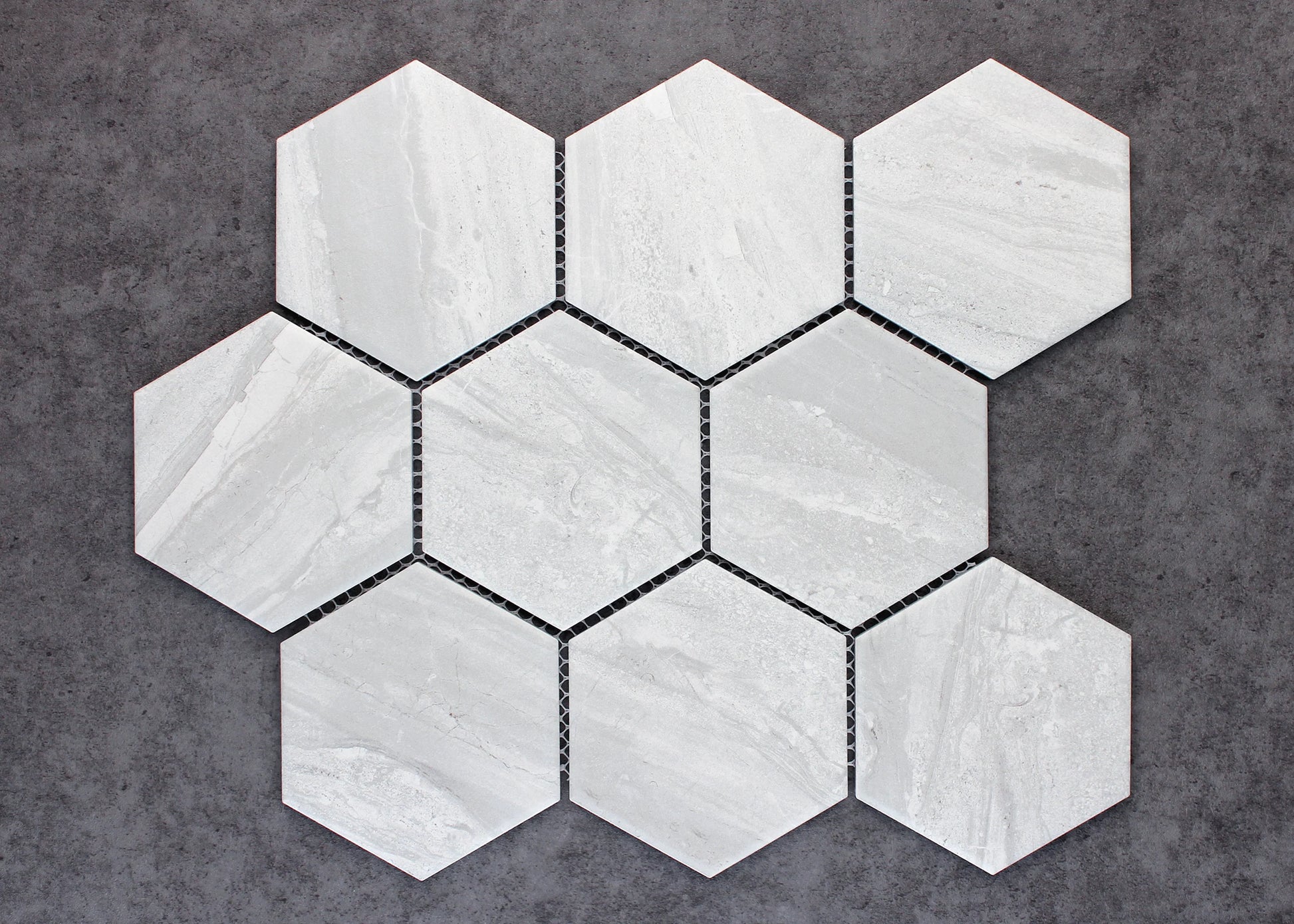 Pack shot of Wooden Grey Matte Porcelain Glazed Hexagon 95x110mm tiles arranged in a seven-piece mesh-backed mosaic sheet, showing soft grey tones and subtle linear stone veining.